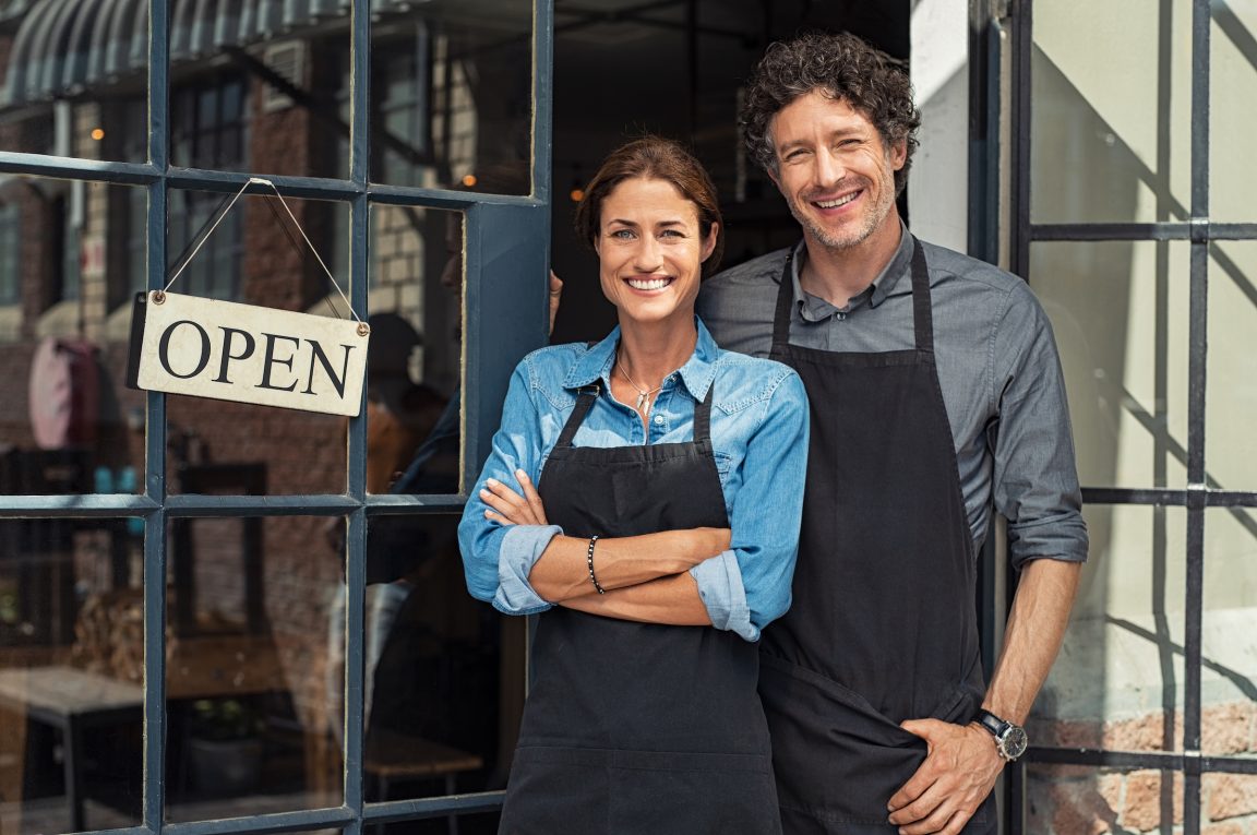two cheerful small business owners smiling and looking at camera while standing at entrance door. happy mature man and mid woman at entrance of newly opened restaurant with open sign board. smiling couple welcoming customers to small business shop