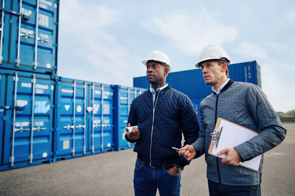 two engineers wearing hardhats and discussing logistics together while standing in a freight yard full of shipping containers