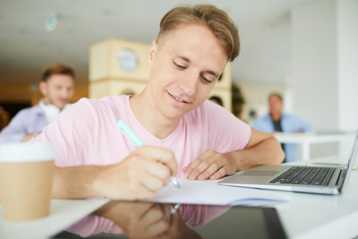 positive confident young guy sitting at desk in library and making notes in workbook while preparing scientific research project